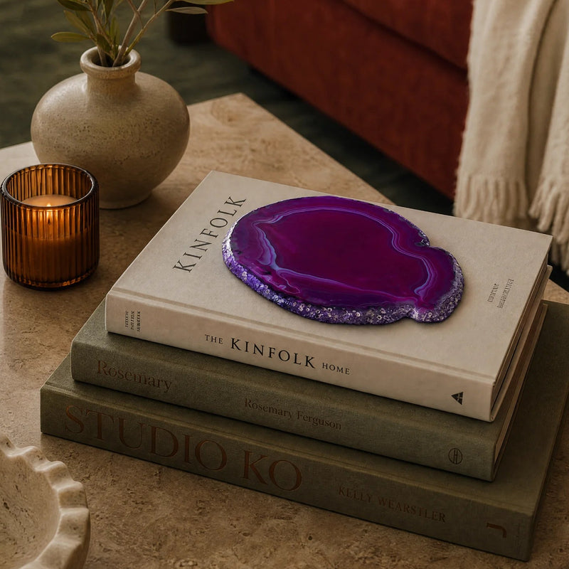 A decorative purple agate slice coaster or tray with a glittery silver edge, displayed on a stack of books on a stone table in a cozy living room setting.
