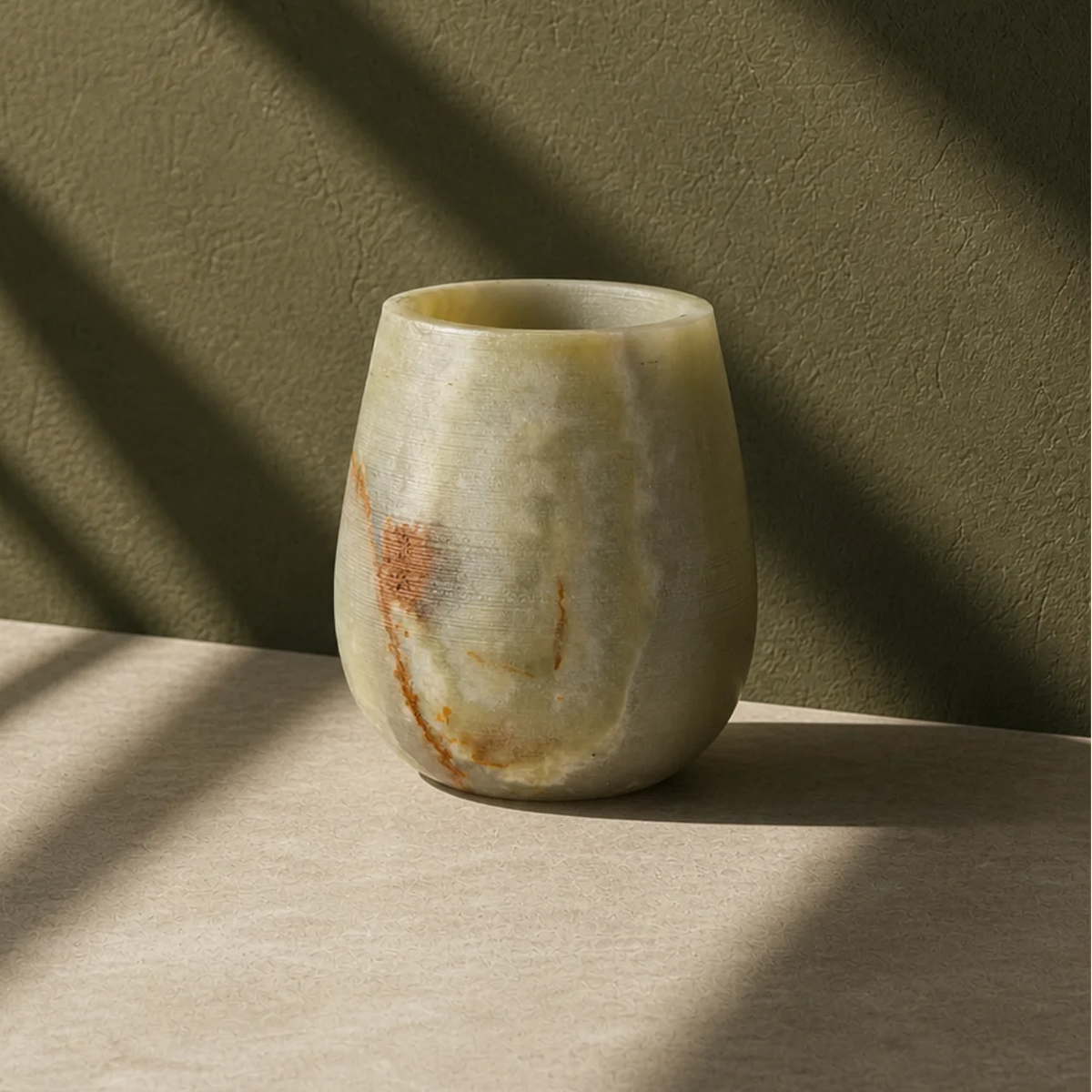 A small rounded stone vase with a natural marbled finish in beige, cream, and brown tones, standing on a light surface against a textured olive-green wall with dramatic diagonal shadows.