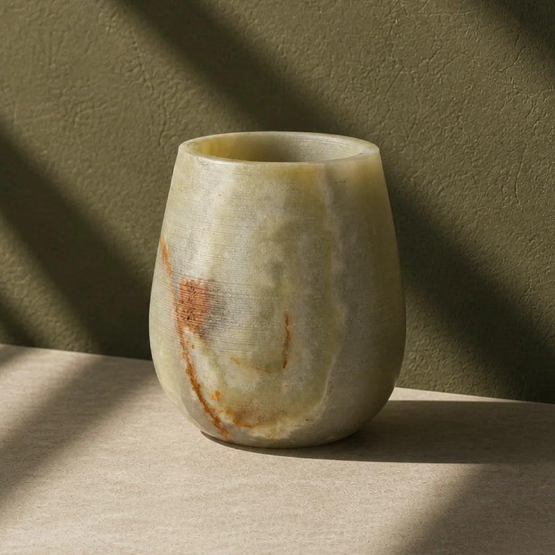 A small rounded stone vase with a natural marbled finish in beige, cream, and brown tones, standing on a light surface against a textured olive-green wall with dramatic diagonal shadows.