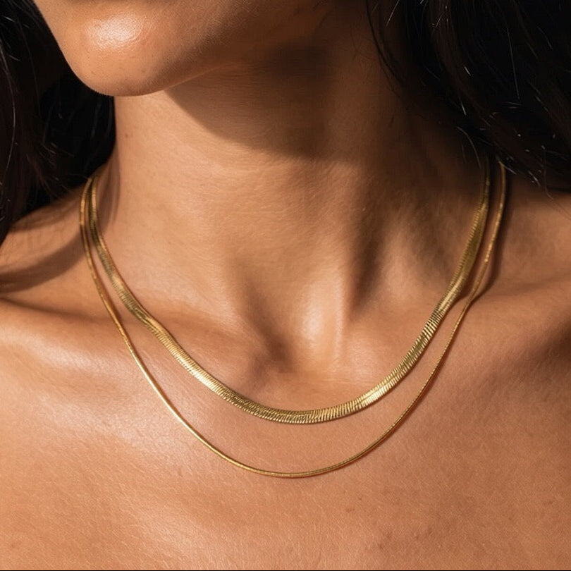 A close-up of a model wearing a delicate double-layered gold chain necklace, showing the fine texture of the chains against her skin.
Gold herringbone and snake chain necklace layered at the collarbone, catching afternoon light against bare skin.