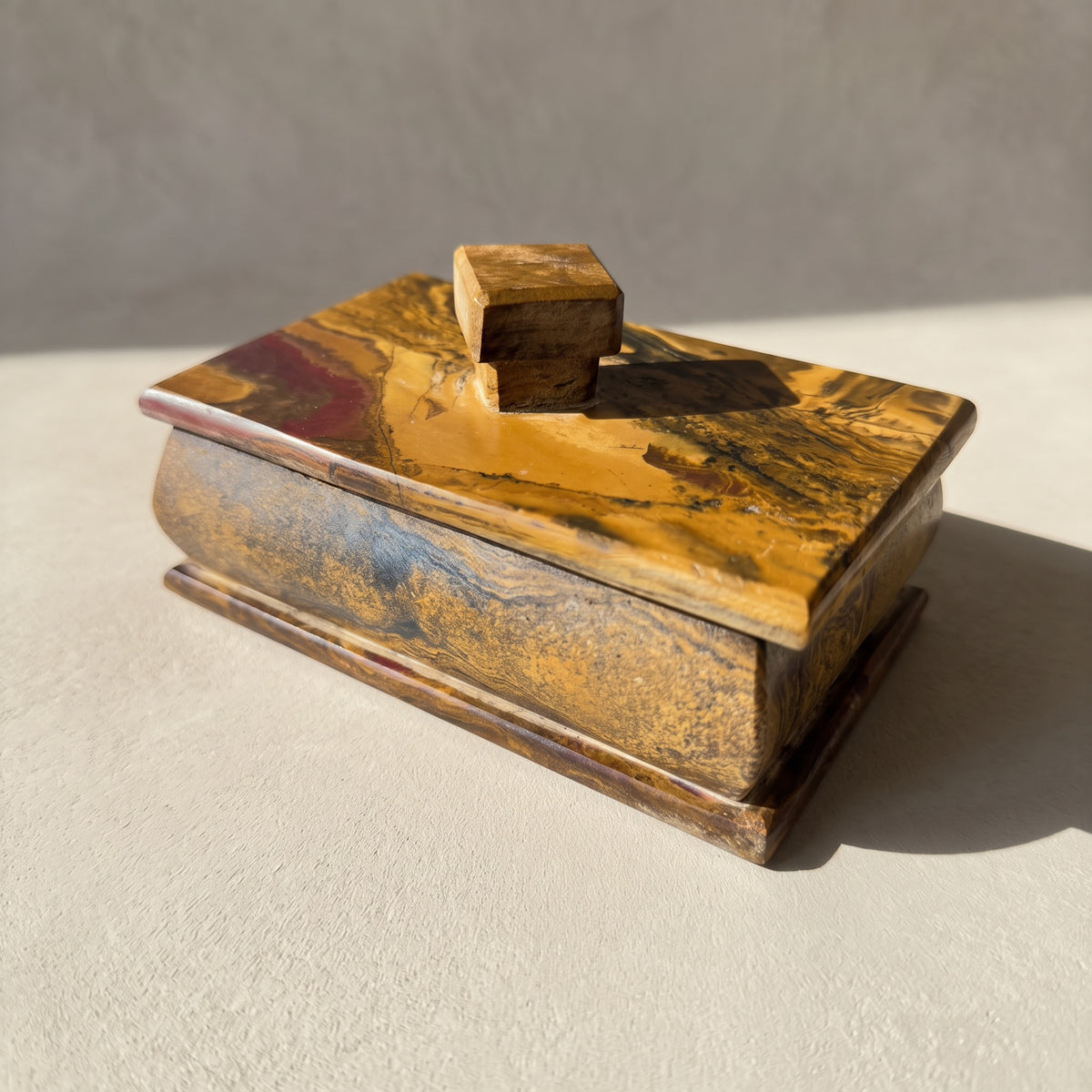 Rectangular onyx keepsake box with lid, three-quarter front view in raking sun on pale plaster, a golden ochre lid with a wine-red corner and dark streaks, a speckled golden brown body and a square two-tier stone knob