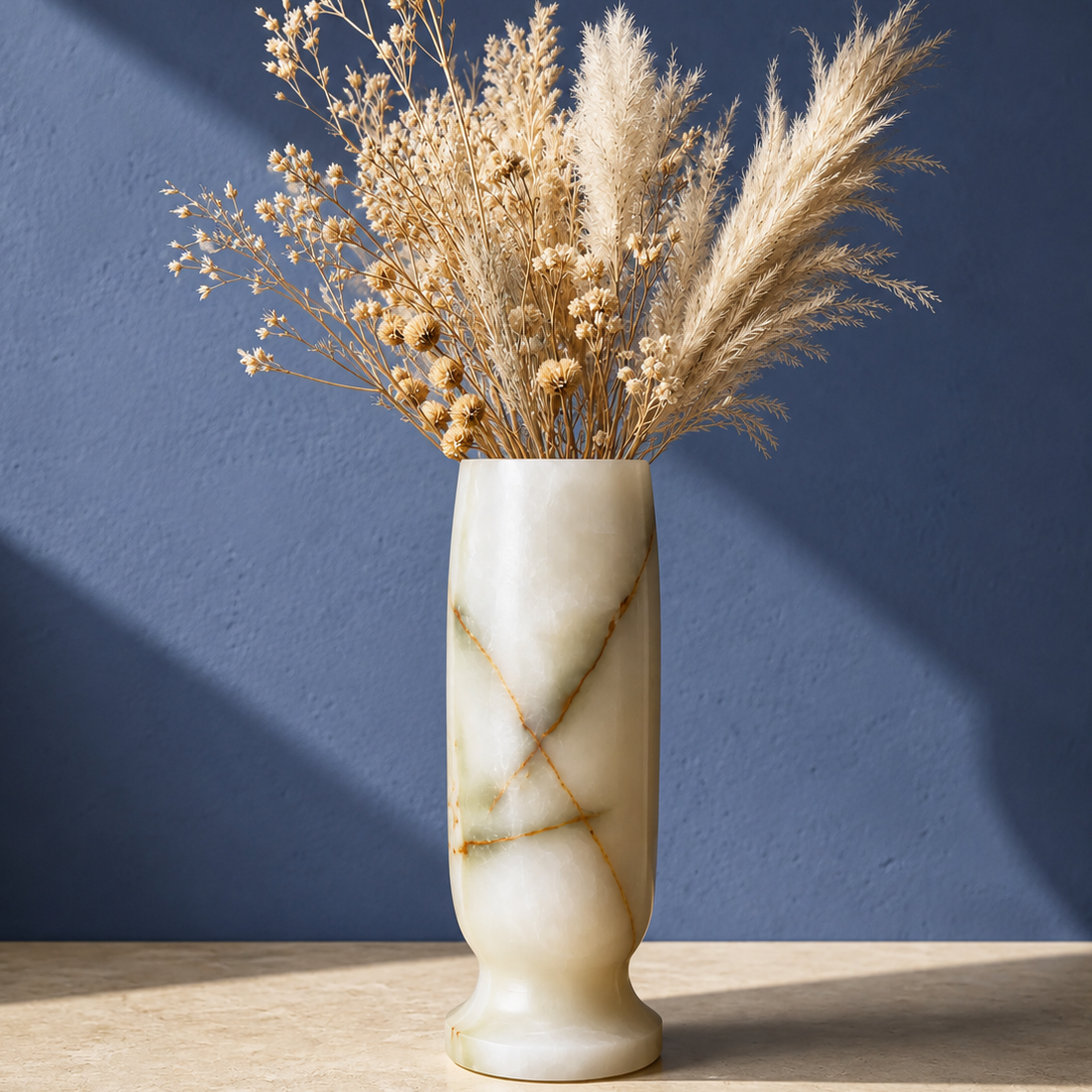 A cream-colored marble green onyx vase with gold veining, filled with dried pampas grass and baby's breath, set on a light wood surface against a textured blue wall.