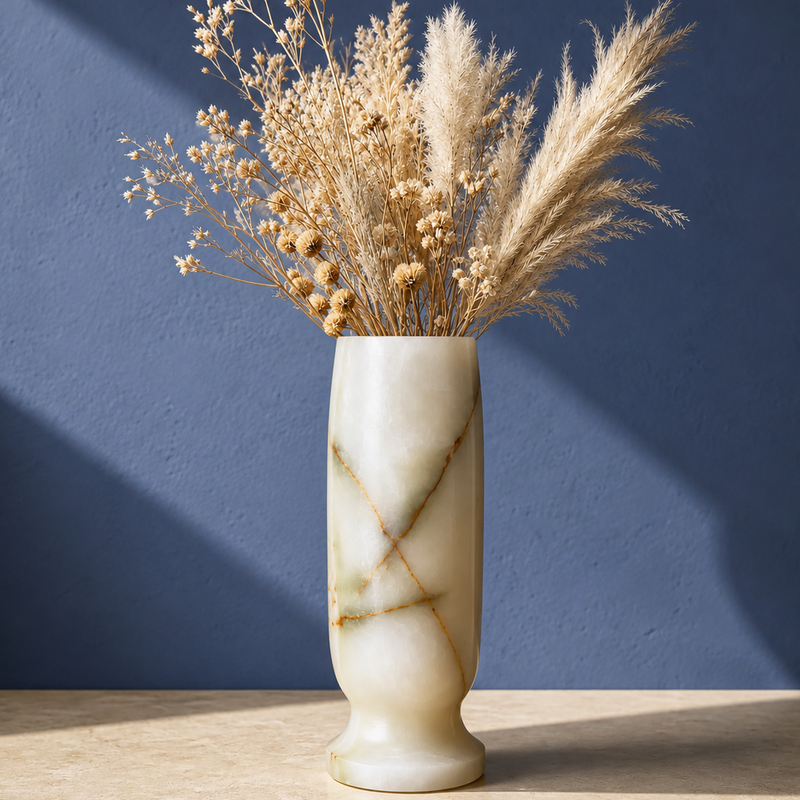 A cream-colored marble green onyx vase with gold veining, filled with dried pampas grass and baby's breath, set on a light wood surface against a textured blue wall.