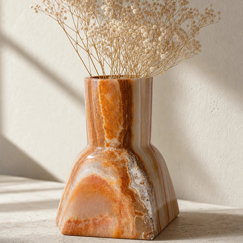 A sculptural ceramic vase made from polished orange and cream agate or onyx stone, holding dried baby's breath flowers, set on a light stone surface against a textured beige wall with soft window shadows.