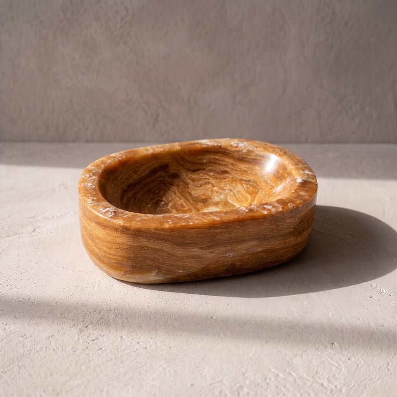 Honey and caramel polished onyx oval catchall bowl, three-quarter view on pale plaster in raking sun