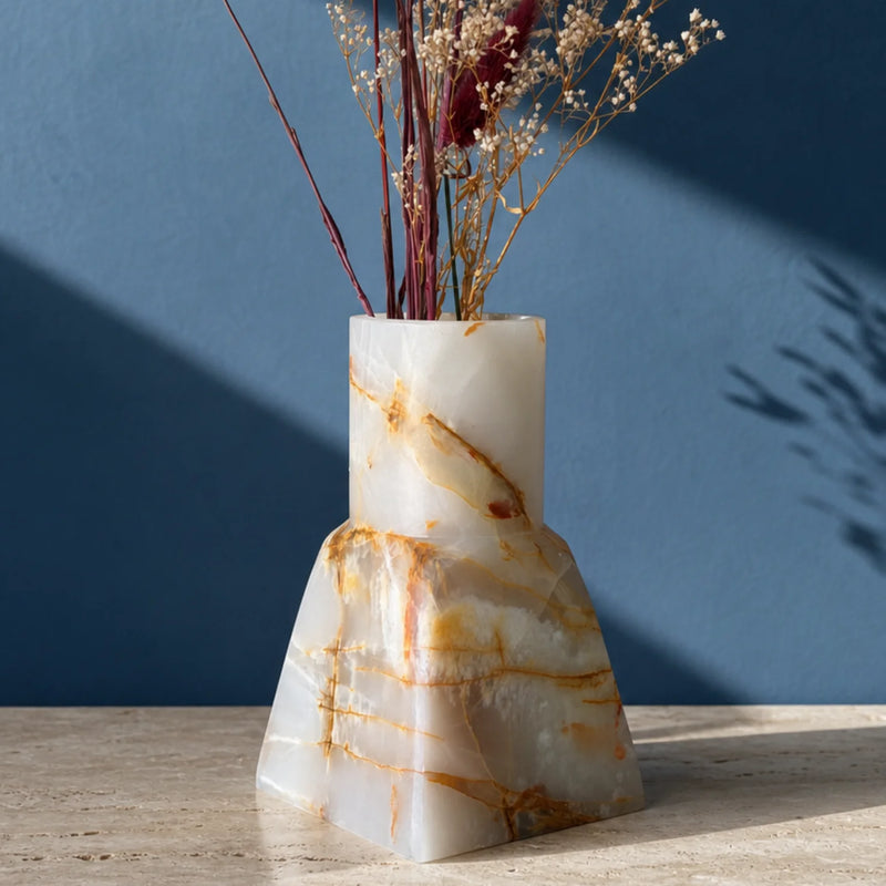 A sculptural white and gold onyx stone vase filled with dried decorative grasses, set on a light stone surface against a deep blue wall with diagonal shadows.