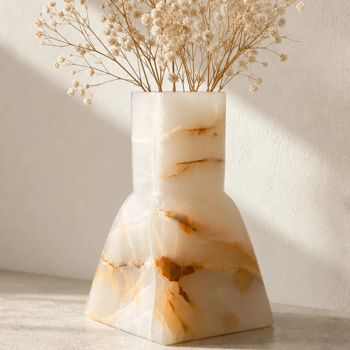 A sculptural vase made of warm-toned marbled stone in cream, beige, and golden brown, holding dried baby's breath flowers, set on a light stone surface against a textured wall with soft diagonal shadows.
