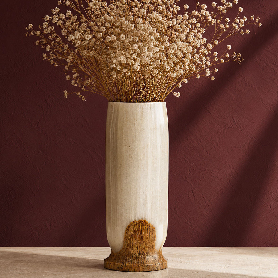 A tall cylindrical ceramic white onyx vase with a matte cream finish and a natural wood-grain textured base, filled with dried baby's breath flowers, set against a dark reddish-brown textured wall.