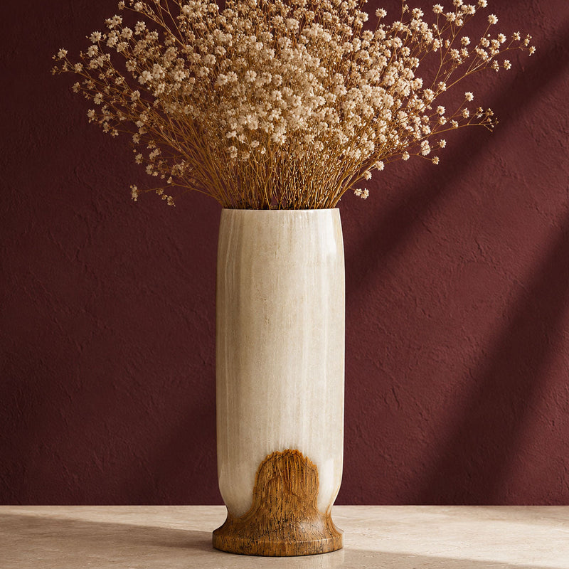 A tall cylindrical ceramic white onyx vase with a matte cream finish and a natural wood-grain textured base, filled with dried baby's breath flowers, set against a dark reddish-brown textured wall.