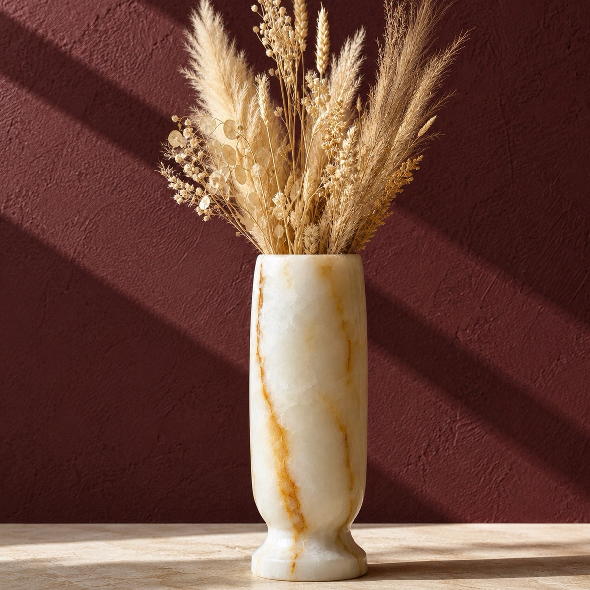 A cream-colored marble onyx vase with natural brown veining, filled with dried wheat and pampas grass, set on a light wooden surface against a textured dark reddish-brown wall with diagonal light shadows.