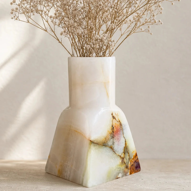 A modern geometric onyx stone vase with dried baby's breath flowers, set on a light beige surface against a textured beige wall with soft leaf shadows.