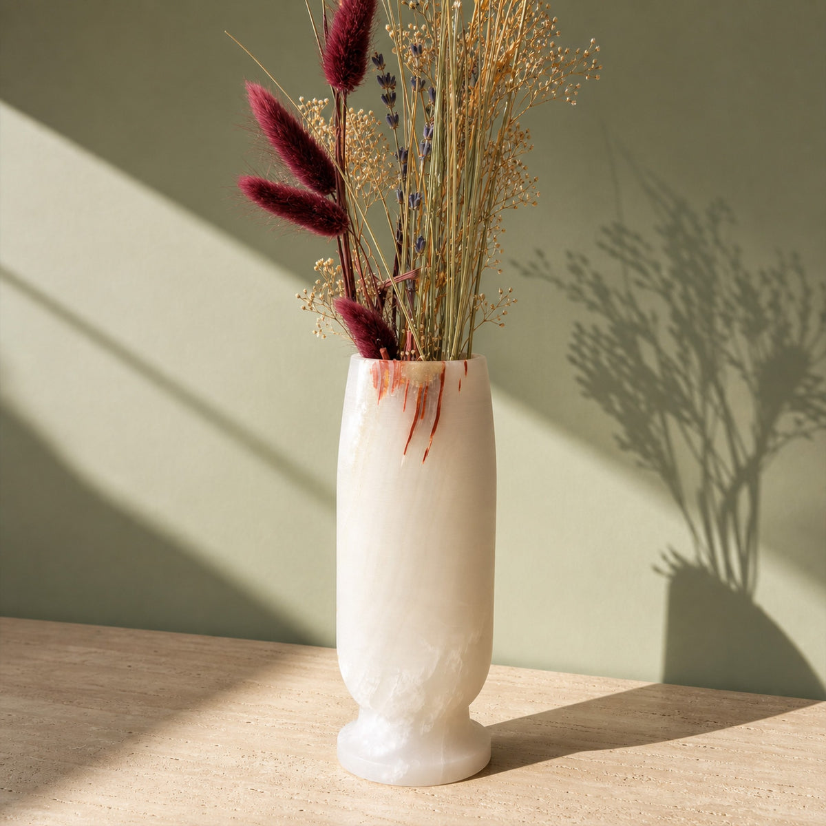 Ivory onyx vase with thin rust streaks running down from the rim and a honey patch at the lip, holding dried burgundy bunny tails, lavender and gypsophila on a travertine table against a sage-green wall