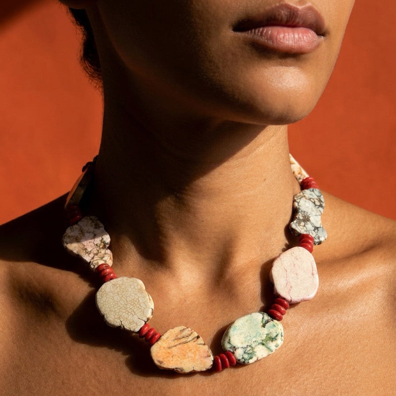 A close-up of a model wearing a chunky statement necklace made of irregular natural stone slices separated by red cord, set against a warm orange background.