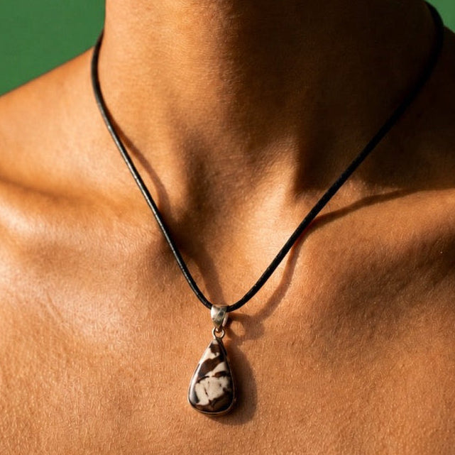 A close-up of a model's neck and upper chest wearing a black cord necklace with a teardrop-shaped carved jasper pendant on a green background.