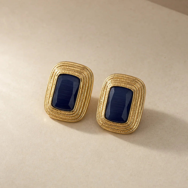 Pair of gold stud earrings with faceted navy cat's eye stones set in stepped, textured gold frames, on a warm beige background.