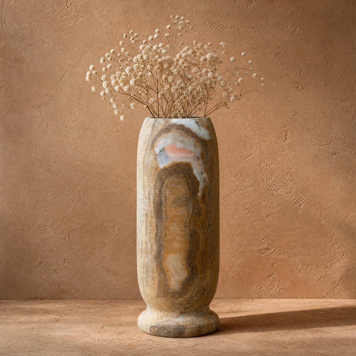 Hand-carved onyx stem vase in ochre, smoke and blush, front view, an amber banded core under a white and blush seam at the rim, holding dried baby's breath against a textured clay-colored wall