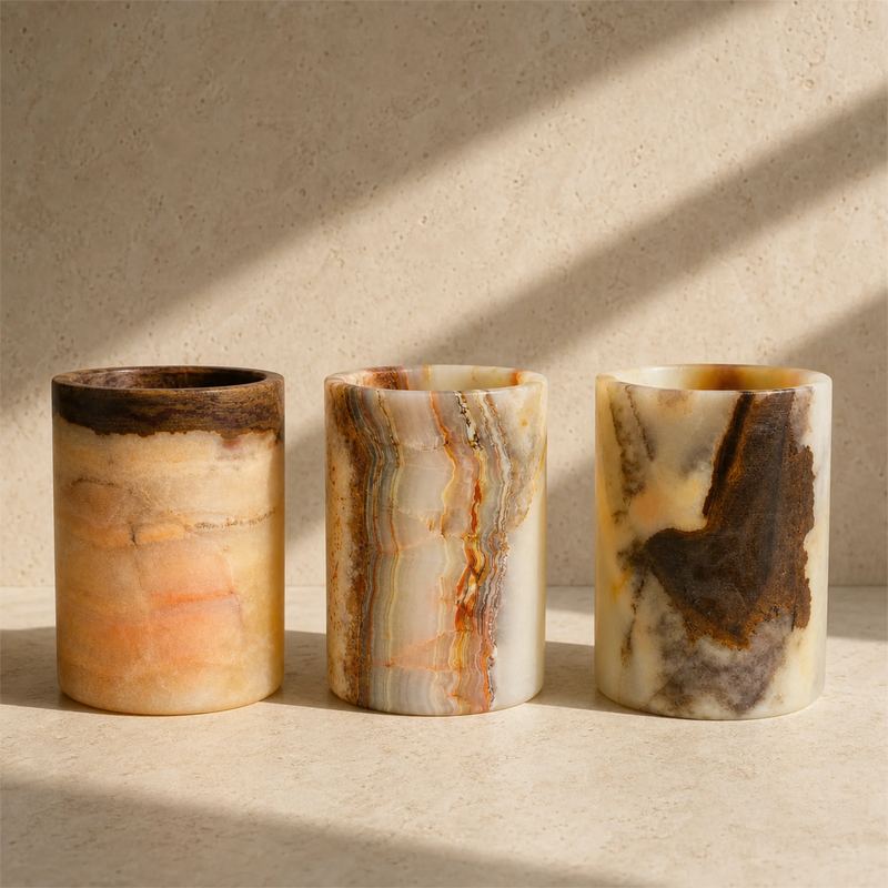 Three cylindrical stone toothbrush holders with natural marbled patterns in beige, cream, orange, and dark brown, arranged on a light stone surface with soft window shadows.
