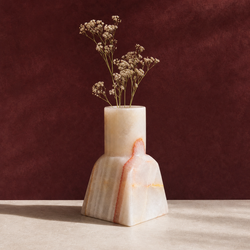 Hand-carved milk-white onyx dune vase with a straight neck on a tapered base, a blush-rust vein arching through the stone, holding dried baby's breath against a deep burgundy wall