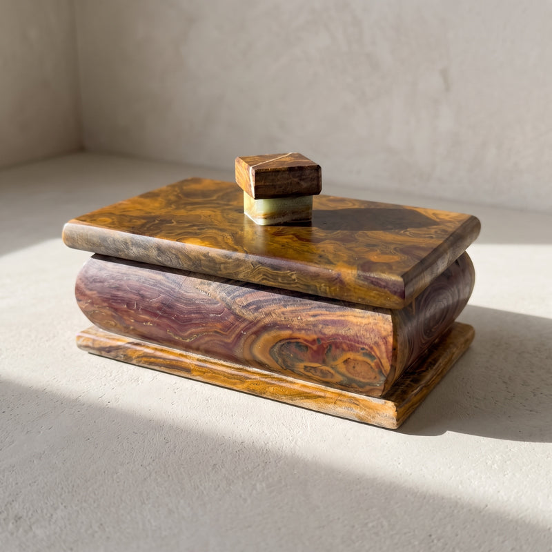 Three-quarter view of an onyx lidded box on pale plaster, its bulged side banded in plum, wine and amber with an olive-gold eye, under a golden caramel lid