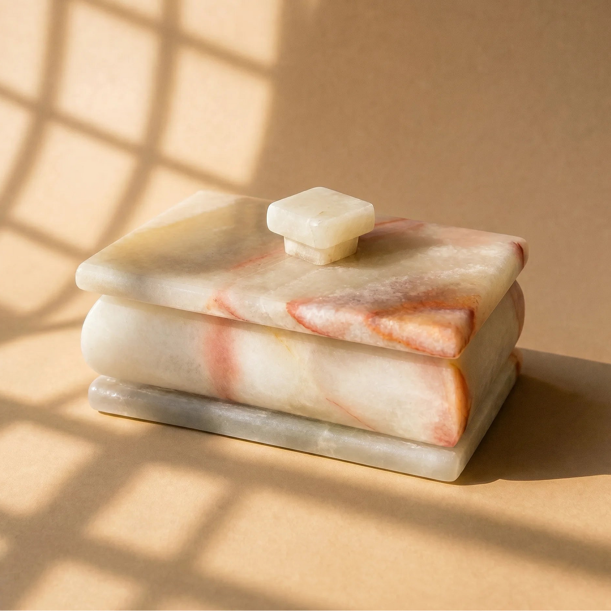 Angled view of ivory onyx lidded box with blush and coral veining and pale square knob in window light