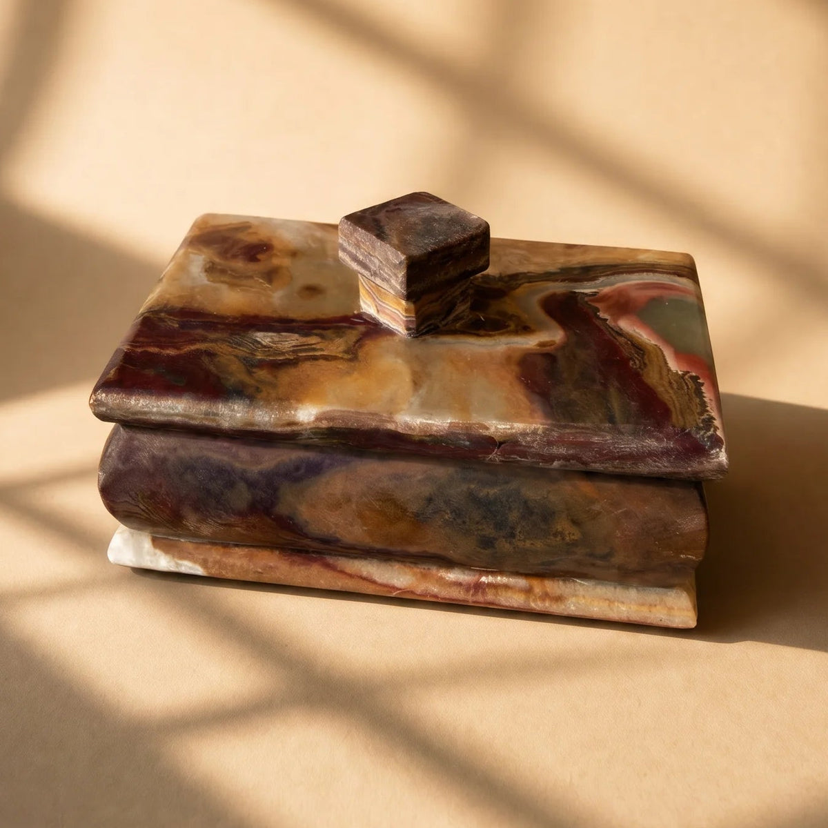 A small rectangular lidded box made of marbled stone in earthy brown, beige, and purple tones, photographed on a warm beige surface with soft window-shadow patterns cast across the scene.