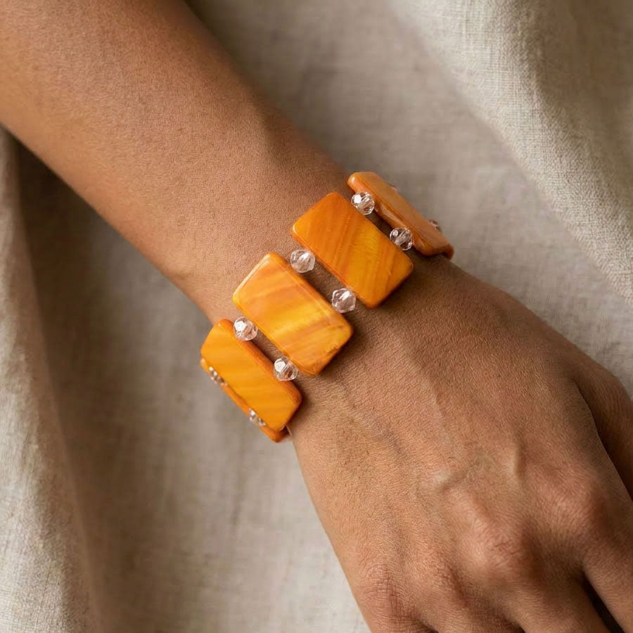 A close-up of a person's wrist and hand wearing a multi-strand bracelet made of warm orange-yellow rectangular stone-like pieces separated by small silver beads, set against a soft beige fabric background.
Orange dyed shell pearl stretch bracelet with faceted clear crystal spacers, worn on the wrist against neutral linen