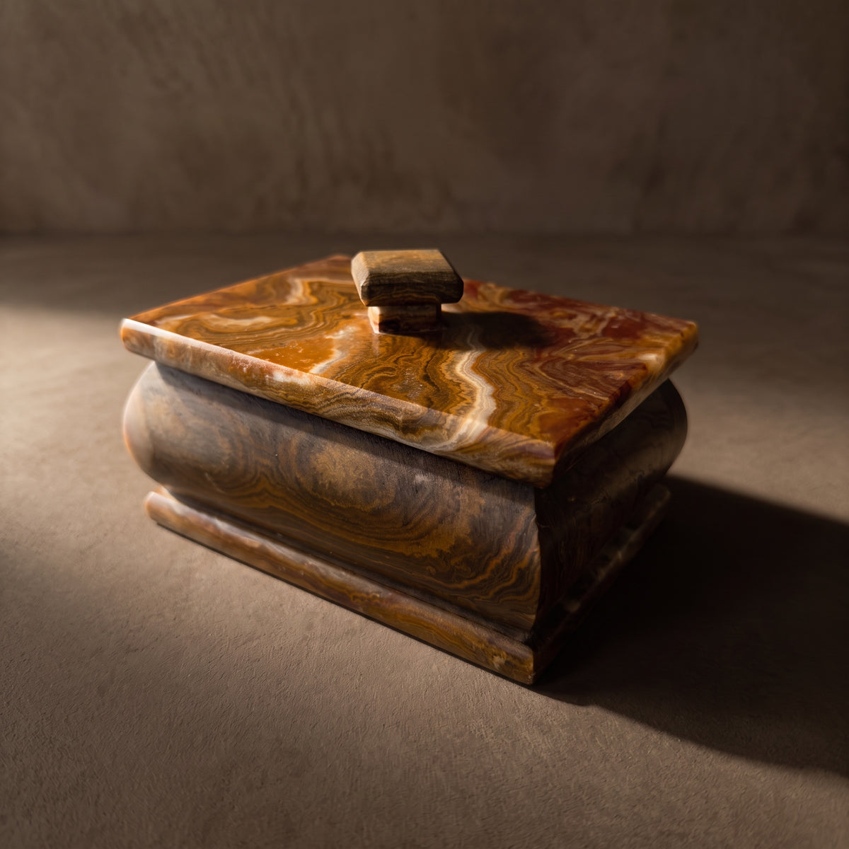 Hand-carved onyx keepsake box with lid in a three-quarter front view: amber, oxblood and cream swirls across the lid, a walnut-brown bulged body and a stepped base, in raking light on warm plaster