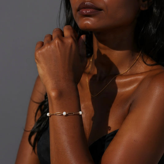 Gold paperclip chain bracelet with pearls shown on a woman’s wrist in natural sunlight.
Pearl and gold paperclip bracelet styled on a woman with deep skin tone, photographed in warm sunlight.