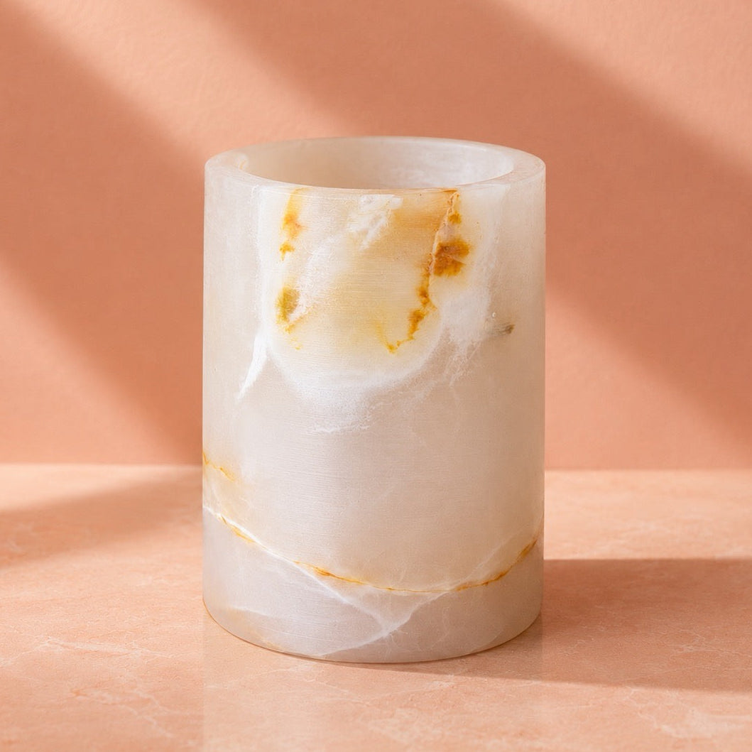 A cylindrical stone candle holder or vase made of milky white onyx with natural golden-brown veining, set on a peach-colored surface against a matching peach background with soft diagonal shadows.