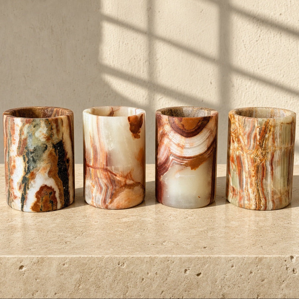 Four cylindrical rainbow onyx stone vessels with marbled patterns in earthy tones of brown, beige, white, and green, lined up on a matching stone surface against a textured wall with soft window shadow patterns. Four one-of-one rust, burgundy, and olive Balochistan onyx cylinder vessels lined up together, one showing a rare black-green mineral vein.