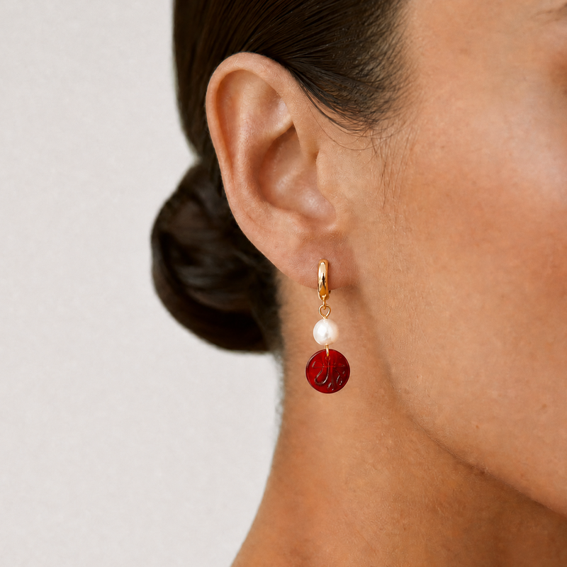 Close-up of a person wearing a red and gold hoop earring with an Allah sign on it on a plain background - Allah jewelry - Mecca beads - red garnet beads