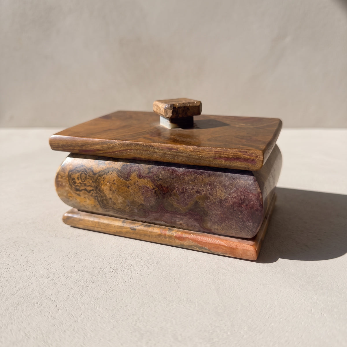 Rust and violet onyx keepsake box with lid in a three-quarter front view: a bulged body clouded in violet, honey gold and rust beneath a brown lid, a stepped base and a small square stone knob, in soft daylight