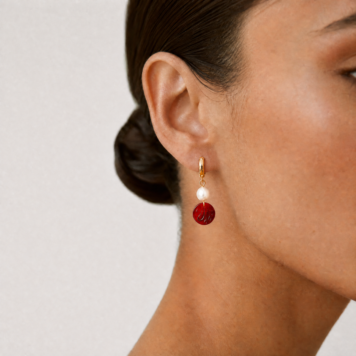 Close-up of a person wearing a red and gold hoop earring with an Allah sign on it on a plain background - Allah jewelry - Mecca beads - red garnet beads