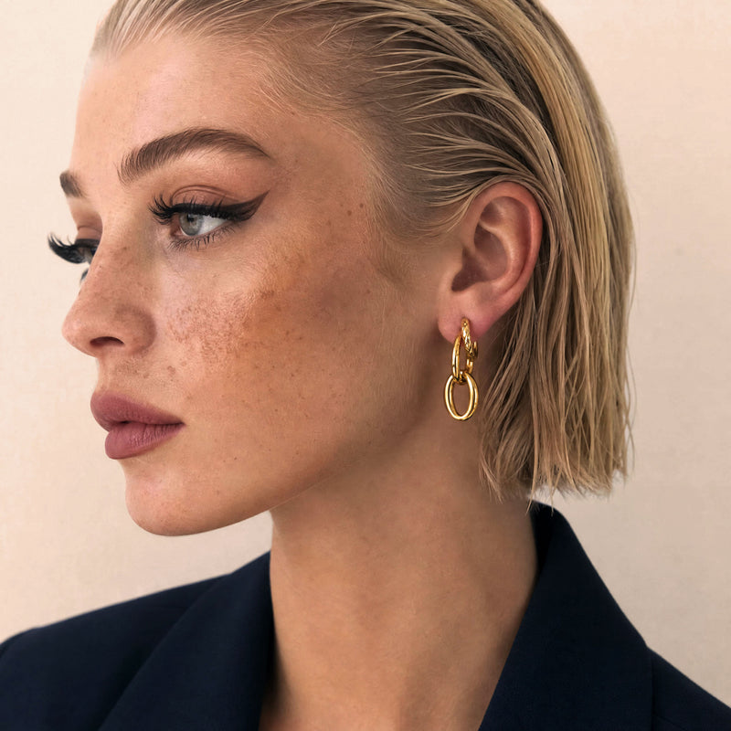 Model in a black blazer wearing a gold double-hoop earring, side profile against a warm neutral backdrop