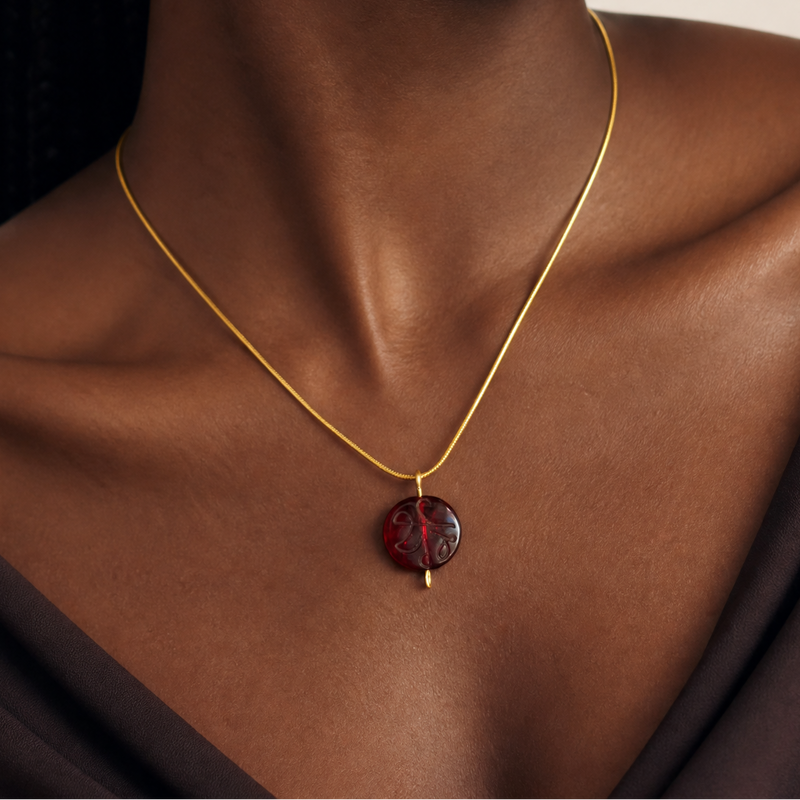 Woman wearing a gold necklace with a red garnet glass pendant against a neutral background