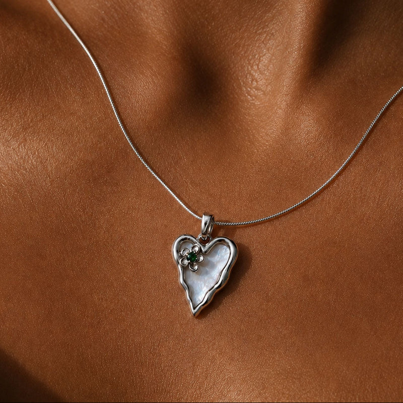 A close-up of a delicate silver chain necklace worn on a model's neck, featuring a heart-shaped pendant with a white mother-of-pearl center and a small clear gemstone.
Silver molten-edge heart pendant with white shell pearl and green cubic zirconia flower, worn on a fine snake chain against sunlit skin at the collarbone