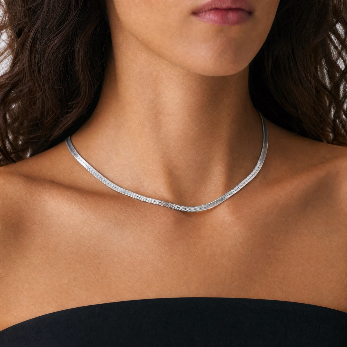 Model wearing a flat silver herringbone chain necklace at the collarbone against neutral backdrop
