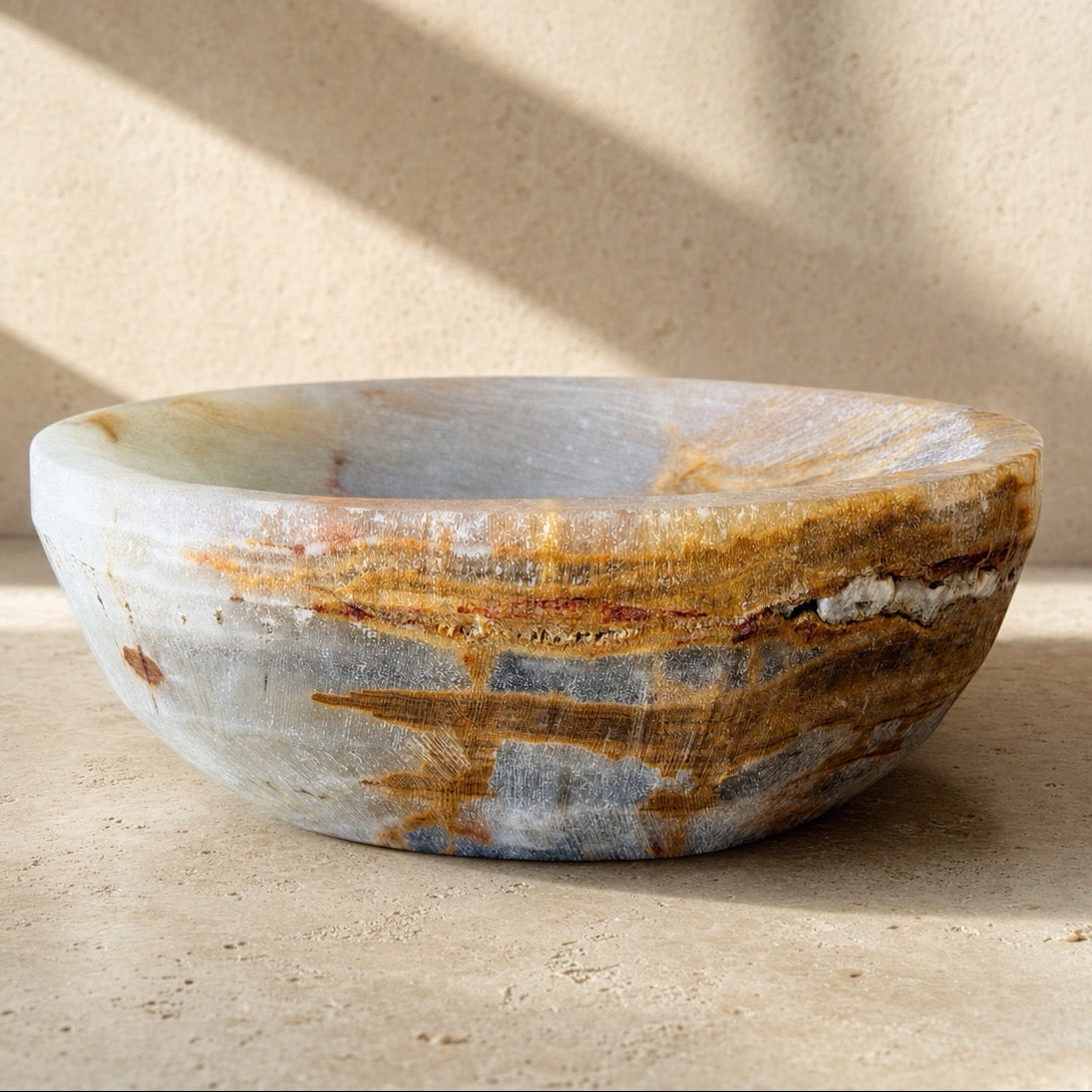 A round stone vessel bowl with natural marbled patterns in gray, beige, and brown, set against a textured beige wall with soft diagonal shadows.
Side profile of a slate grey onyx catchall bowl with an amber and rust vein sweeping across the surface, set against a sunlit stone wall.