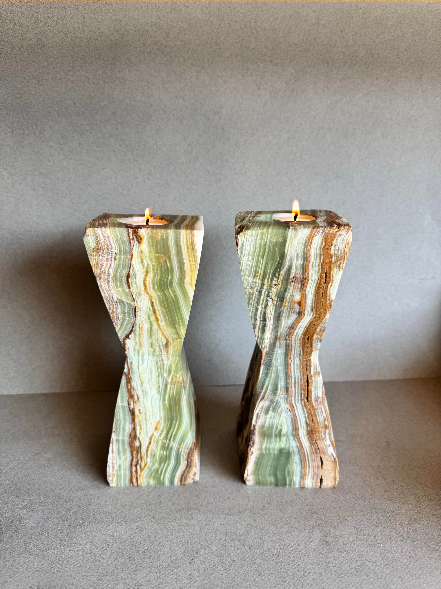 Pair of hand-carved green onyx hourglass candle holders with lit tealight candles, showcasing warm natural veining in sage, clay, and honey tones against a neutral background.