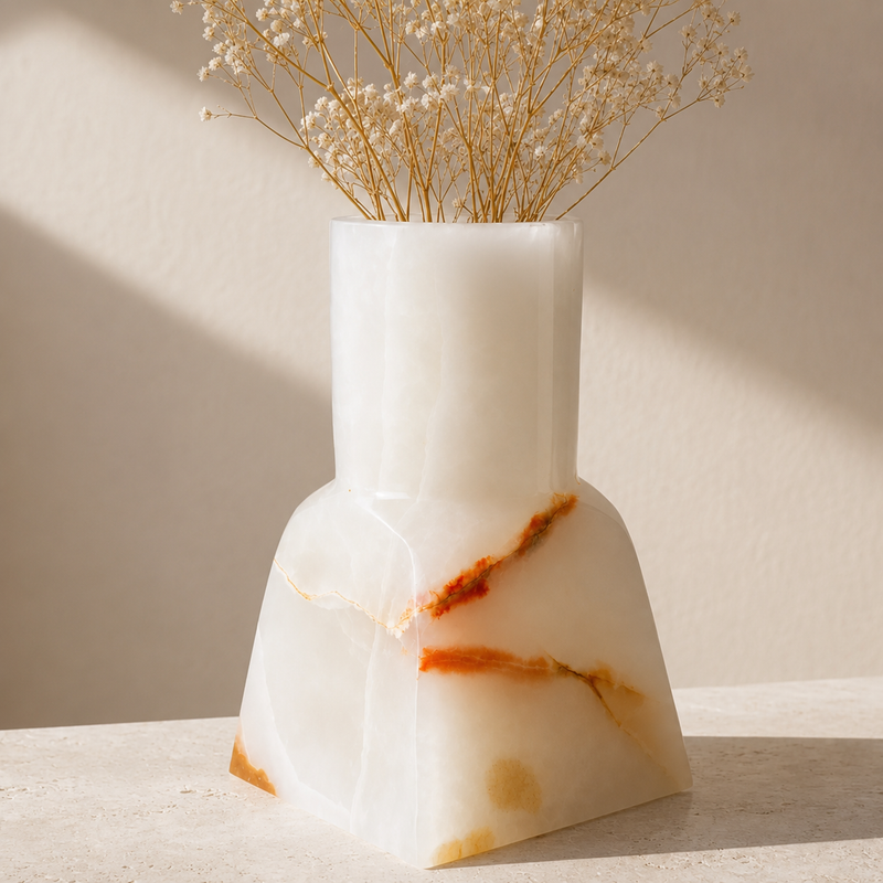 A modern white onyx stone vase with natural orange-brown veining, holding dried baby's breath flowers, set on a light stone surface against a beige wall with soft diagonal shadows.