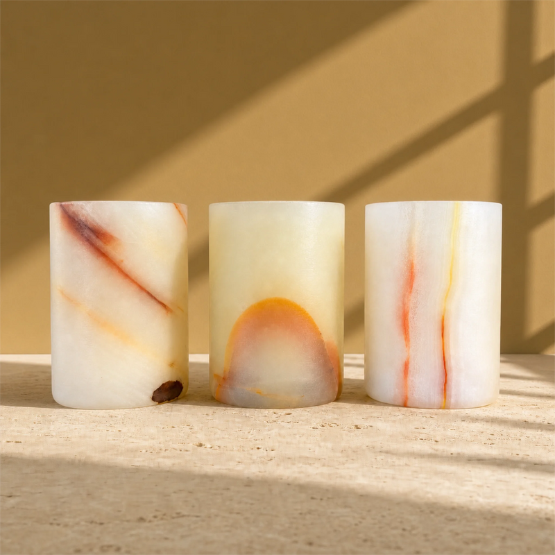Three cylindrical stone candle holders with marbled white, orange, and brown patterns, arranged on a light stone surface against a warm beige background with soft window shadow patterns.