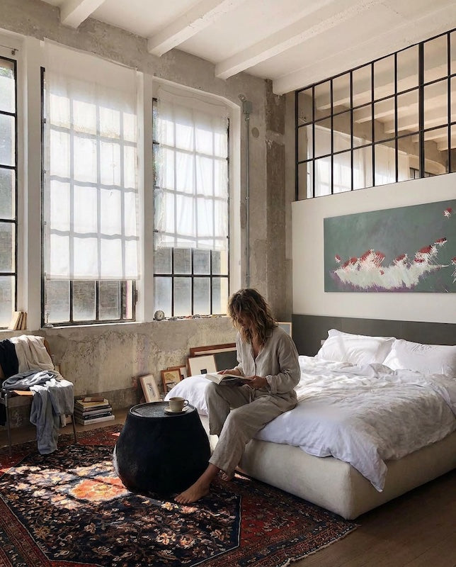 A woman reads on an unmade bed in a sunlit concrete loft, books stacked on the floor, art leaning on the wall, a worn rug at her feet