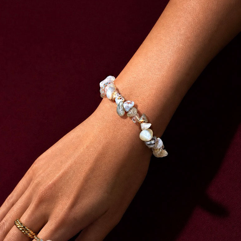 A close-up of a person's wrist wearing a bracelet made of irregular white and gray shell-like beads, against a dark burgundy background.