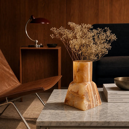 A sculptural hand-carved onyx vase in amber and cream marbling on a marble coffee table in a styled maximalist living room, holding dried flowers.