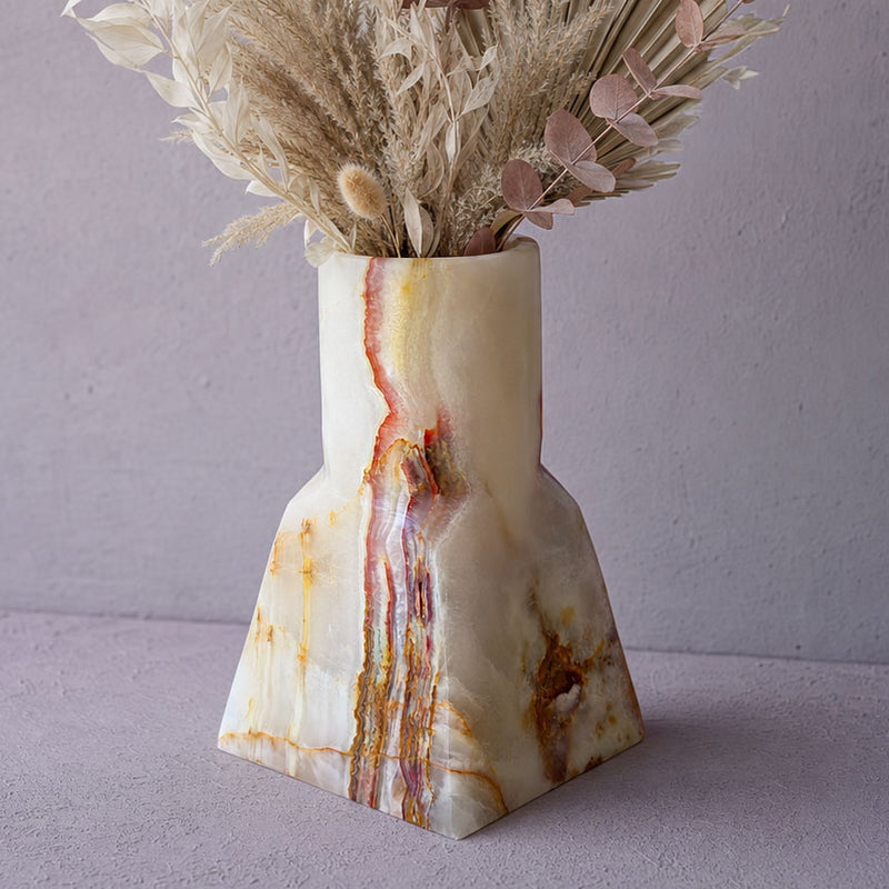 A sculptural onyx-style vase with dried botanical stems, set against a soft lavender-gray background.