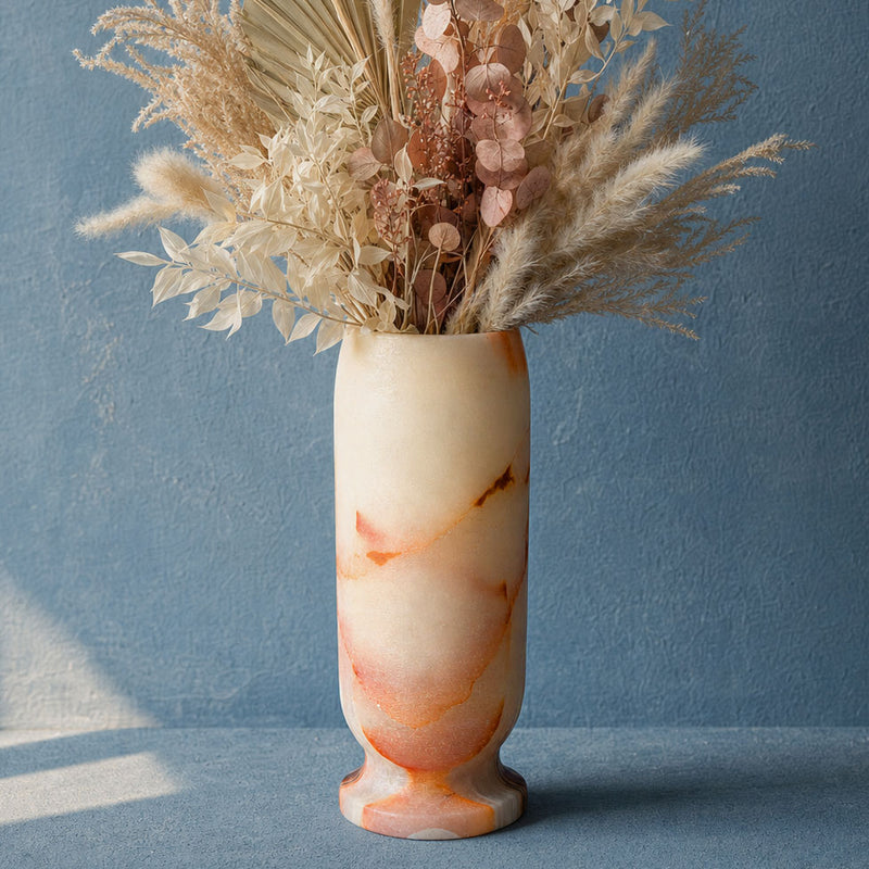 A cream and peach onyx marble vase filled with dried pampas grass and pink flowers, set against a textured blue wall.