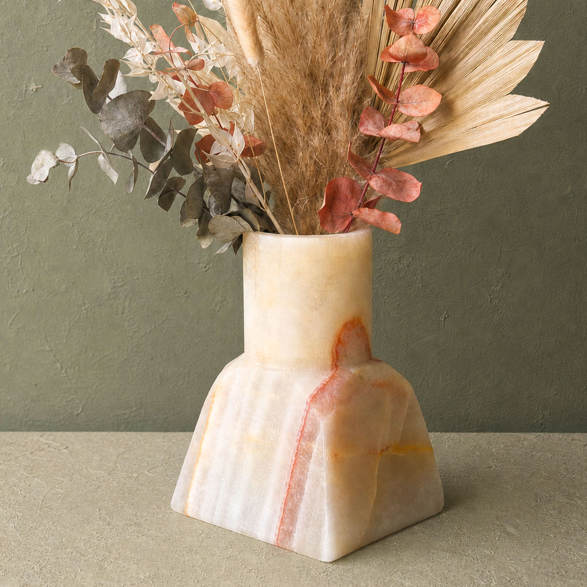 A sculptural, tapered onyx stone vase made of cream and peach marble with a vertical pinkish-red veining, filled with dried botanicals, set against a textured olive green wall.