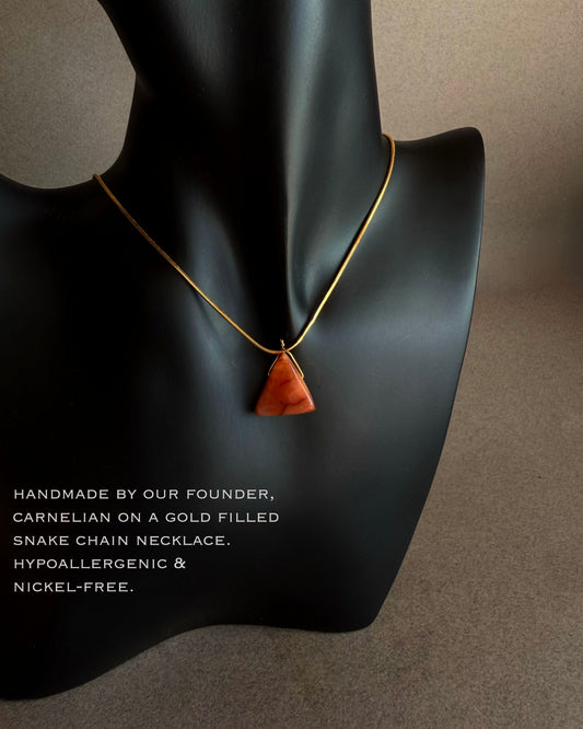 Necklace with a triangular pendant on a mannequin bust against a neutral background Close up of carnelian triangle gemstone pendant with warm rust and terracotta tones
