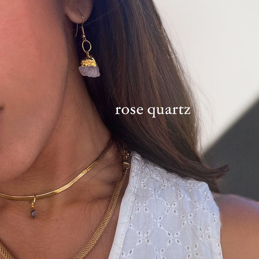 Close-up of a person wearing gold earrings with rose quartz stones against a neutral background.