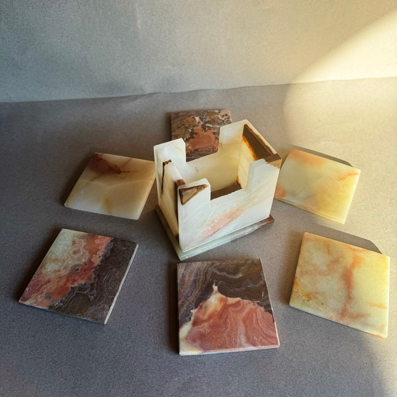 Set of square stone coasters with matching holder, hand-carved from onyx in ivory, honey, and rust tones, shown scattered on table.